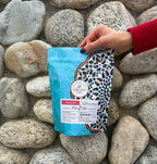 A hand wearing a red sweater holding a 12oz bag of Noosh Beans Coffee Shiraz Signature Blend outdoors against a backdrop of smooth, natural river rocks. The coffee bag features a light blue honeycomb pattern on the left and an intricate, colorful mosaic design on the right. The white label displays the Noosh Beans logo, a 70% Arabica and 30% Robusta ratio, and medium-dark roast tasting notes.